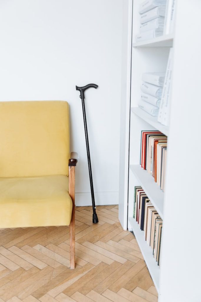 A bright reading nook featuring a yellow armchair, a black cane, and a bookcase filled with books.